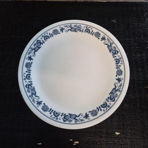 Puede incluir: Plato de cerámica blanco con un diseño de borde floral azul. El plato presenta un patrón repetitivo de flores y hojas estilizadas en color azul oscuro. El plato redondo tiene un diseño simple y clásico, adecuado para el uso diario o ocasiones especiales.