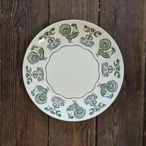May include: A round off-white ceramic plate featuring a scalloped central border and a repeating pattern of dark green roosters and stylised floral motifs arranged around the perimeter of the plate's surface in a symmetrical circular design.