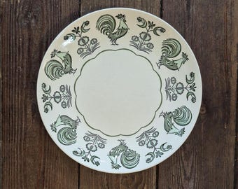 May include: A round off-white ceramic plate featuring a scalloped central border and a repeating pattern of dark green roosters and stylized floral motifs arranged around the perimeter of the plate's surface in a symmetrical circular design.