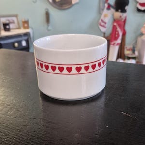 May include: A white ceramic bowl with a red heart pattern around the rim. The bowl is cylindrical and sits on a dark surface. The background is blurred, suggesting a shop or display setting.