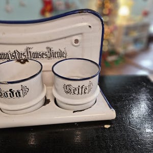 May include: A vintage white enamel soap dish with two cylindrical holders and a blue trim. The holders are labeled "Soda" and "Seife" in black script. The dish has a curved top and is set on a dark surface.