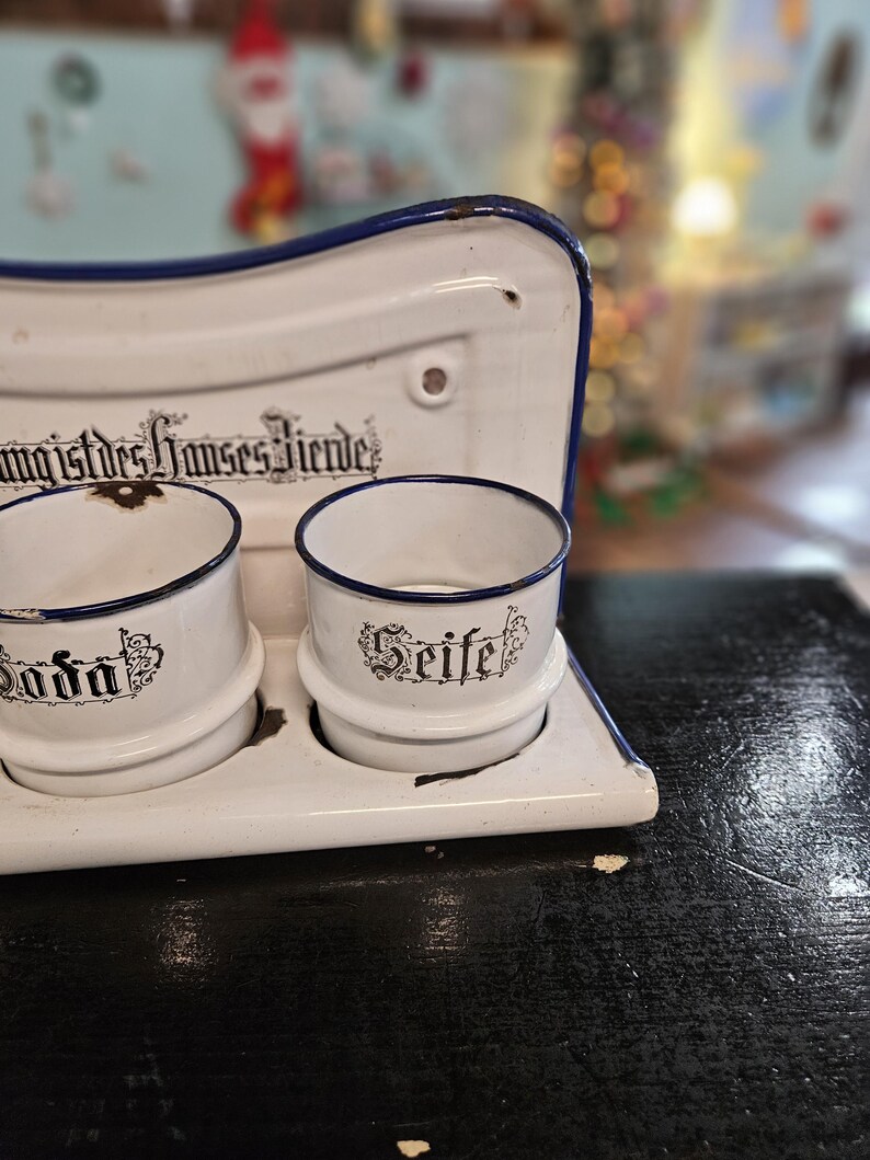 May include: A vintage white enamel soap dish with two cylindrical holders and a blue trim. The holders are labeled "Soda" and "Seife" in black script. The dish has a curved top and is set on a dark surface.