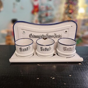 May include: A white enamel kitchen organizer with three small cylindrical containers labeled "Sand", "Soda", and "Seife" in black script. The organizer has a blue trim and a backboard with the text "Ordnung ist des Himmels Freude".