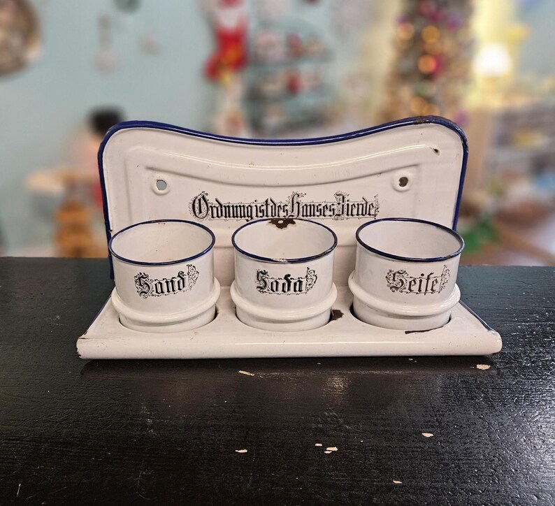 May include: A white enamel kitchen organizer with three small cylindrical containers labeled "Sand", "Soda", and "Seife" in black script. The organizer has a blue trim and a backboard with the text "Ordnung ist des Himmels Freude".
