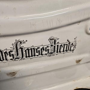 May include: Close-up of a white enamelware pot with a blue rim. The pot has a decorative black inscription in a gothic font. The enamel surface shows signs of wear and tear, with scratches and small imperfections.