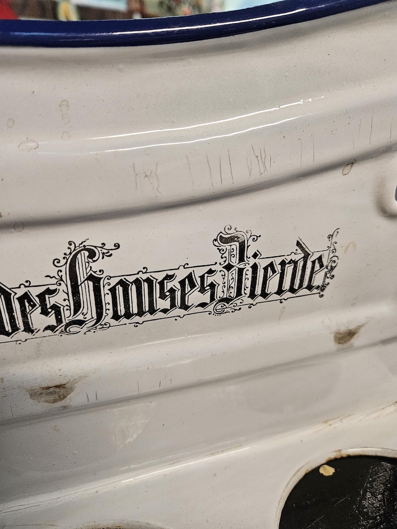May include: Close-up of a white enamelware pot with a blue rim. The pot has a decorative black inscription in a gothic font. The enamel surface shows signs of wear and tear, with scratches and small imperfections.