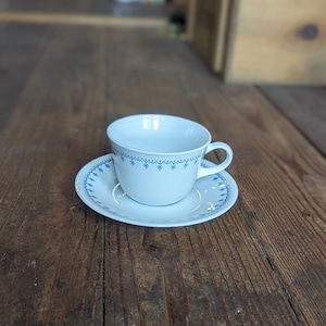 Può includere: Una tazza da tè e un piattino bianchi con un motivo a fiocco di neve blu. La tazza ha un manico e il piattino ha un bordo ondulato.