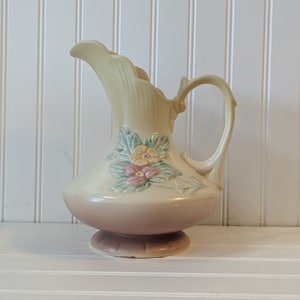 Puede incluir: Jarra de cerámica vintage con cuerpo color crema y base rosa. La jarra presenta un asa y un pico con borde festoneado. Detalles florales en relieve en colores pastel adornan el frente. La jarra mide aproximadamente 20 cm de altura.