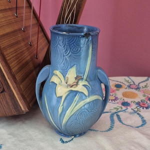 May include: A blue ceramic vase featuring two side handles and a raised relief design of a yellow lily flower. The surface of the vase has a textured, swirling pattern throughout, and there is visible wear along the top rim of the vessel.