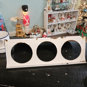May include: A white metal kitchen range top with three circular openings. The range top has a rectangular shape with a slightly distressed appearance. The background includes Christmas decorations and a white ceramic bowl with the word "Santa" on it.