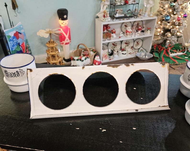 May include: A white metal kitchen range top with three circular openings. The range top has a rectangular shape with a slightly distressed appearance. The background includes Christmas decorations and a white ceramic bowl with the word "Santa" on it.