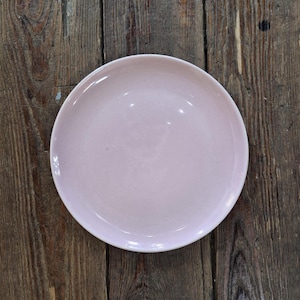 Puede incluir: Un plato redondo de cerámica rosa pálido con un borde ligeramente elevado. El plato tiene un acabado brillante y está sobre una superficie de madera rústica y envejecida. El plato tiene unos 25 cm de diámetro.