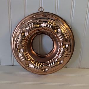 May include: A copper-colored Bundt cake pan with a central hole and decorative embossed patterns. The pan has a circular shape with a hanging loop at the top. The pan is made of metal and has a shiny finish.