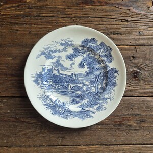 May include: A round white ceramic plate featuring a blue transferware pattern. The design depicts a landscape with a stone bridge over a river, a castle on a hill, and large trees. The illustration is rendered in a detailed, etched style.