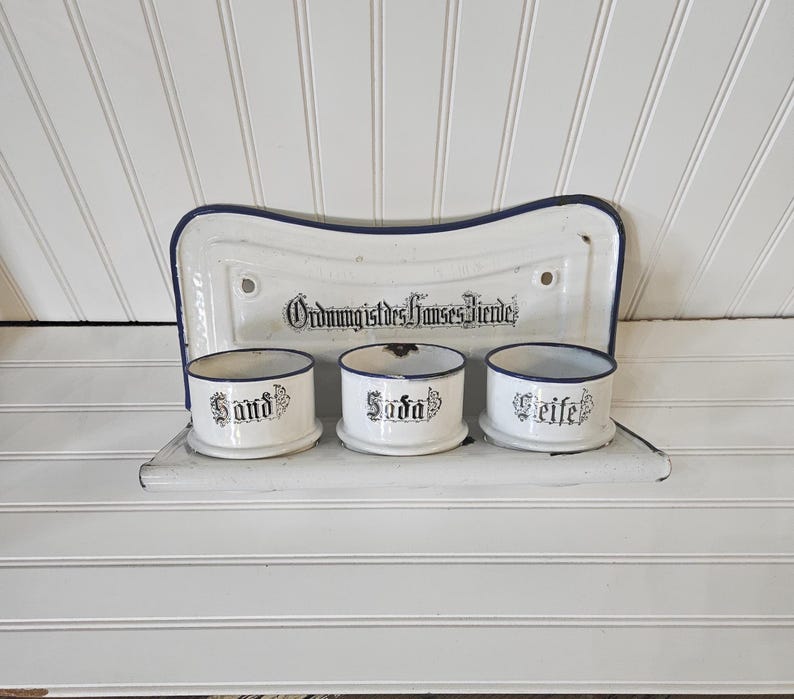 May include: A white enamel wall-mounted shelf with a blue trim, holding three small cylindrical containers. The containers are labeled "Sand", "Soda", and "Seife" in black script. The shelf has the text "Ordnung ist des Hauses Zierde" in black.