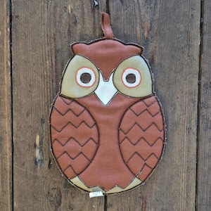 Op de afbeelding: Een bruine en beige uilvormige pannenlap met een lus om op te hangen. De uil heeft grote, ronde ogen met oranje en witte accenten en een witte snavel. Het lichaam heeft een zigzagpatroon in donkerder bruin.