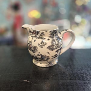May include: A cream-colored ceramic creamer pitcher featuring a black toile pattern. The design depicts various outdoor scenes with figures, trees, and small birds. The pitcher has a rounded body, a curved handle, and a small pouring spout.