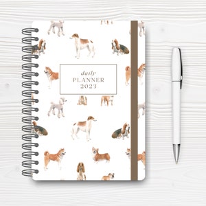 May include: A white daily planner with a brown spine and a repeating pattern of watercolor dogs. The planner has a title that reads "daily planner 2023".