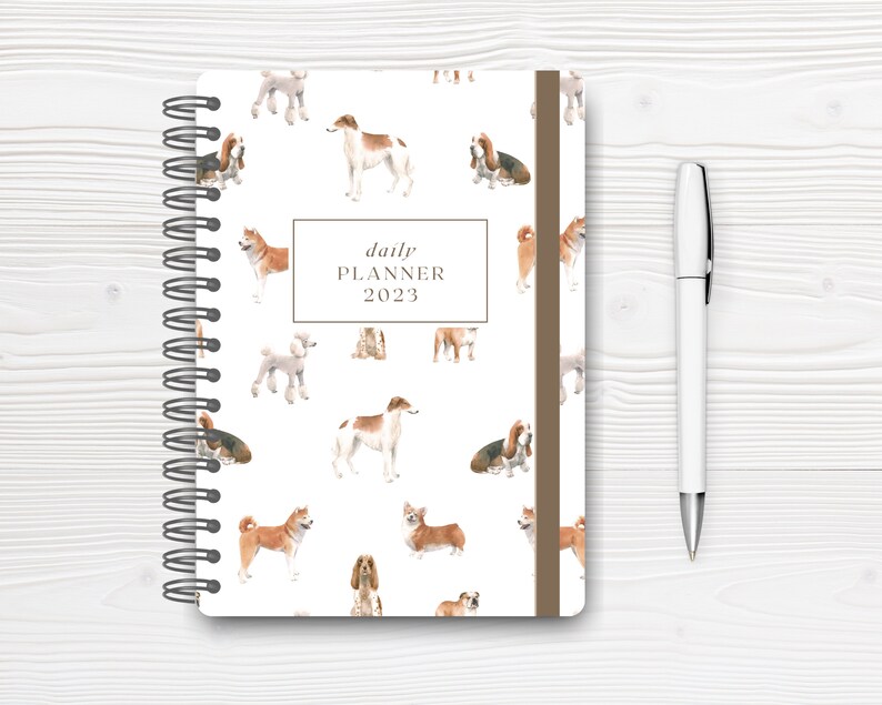 May include: A white daily planner with a brown spine and a repeating pattern of watercolor dogs. The planner has a title that reads "daily planner 2023".