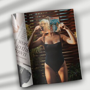 May include: A woman in a black swimsuit is holding a book titled "Capit&atilde;es da Areia" by Jorge Amado in front of her face. The book cover features a red, white, and blue design.