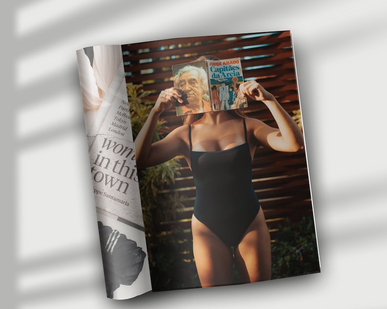 May include: A woman in a black swimsuit is holding a book titled "Capit&atilde;es da Areia" by Jorge Amado in front of her face. The book cover features a red, white, and blue design.