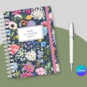 May include: A daily planner with a floral design. The cover features a variety of flowers in shades of pink, blue, white, and yellow on a dark blue background. The planner has a pink and white striped spine and a silver coil binding. The text "daily planner 2023" is printed in white on a white rectangle.