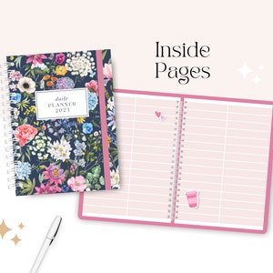 May include: A daily planner with a floral design on the cover. The inside pages are pink with lines for writing. The planner has a pink binding and a silver pen is next to it.