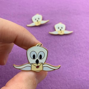 May include: Three gold enamel pins of a white owl with large eyes holding a yellow envelope with a red seal. The owl is flying with its wings outstretched.