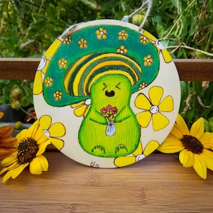 May include: A green mushroom character with a yellow and green striped hat and a bouquet of flowers, painted on a round wooden sign with yellow flowers.