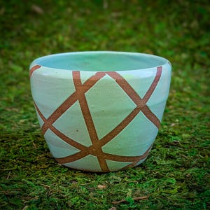 May include: A handmade ceramic bowl with a light teal glaze and a geometric pattern in a warm brown color. The bowl has a rounded shape and is suitable for various uses, such as a planter or decorative piece.