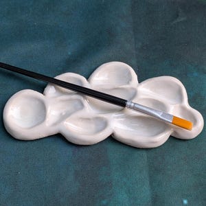 May include: A white ceramic cloud-shaped paint palette with several wells, holding a black paintbrush with a silver ferrule and orange bristles. The palette is on a teal fabric surface.