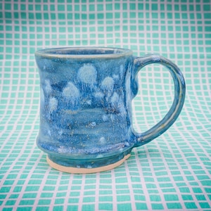 May include: A handmade ceramic mug with a blue glaze. The mug has a rounded body, a sturdy handle, and a light-colored base. The blue glaze has lighter blue accents, creating a textured appearance. The mug is set against a teal and white checkered background.