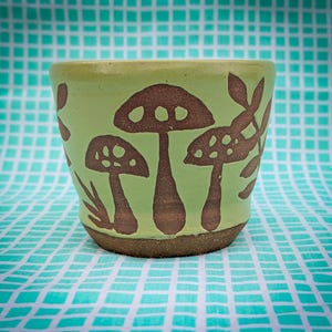 May include: A ceramic mug with a light green background and brown mushroom illustrations. The mug has a rounded shape and a textured, brown base. The mushroom design includes various sizes and shapes, adding a whimsical touch to the handmade pottery.