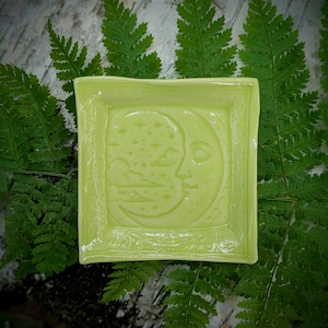 May include: A square, light lime green ceramic trinket dish featuring a relief design of a crescent moon with a face, surrounded by stars and clouds. The dish has a smooth, glossy finish and a slightly raised border with decorative detailing.