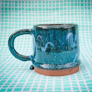 May include: A handmade ceramic mug with a teal glaze and a brown base. The mug has a large handle and a unique speckled pattern. The mug is approximately 4 inches tall and 3.5 inches in diameter.