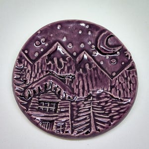 May include: A round, purple ceramic piece featuring a mountain scene with a cabin, trees, and a night sky with a crescent moon and stars. The design is carved into the surface.