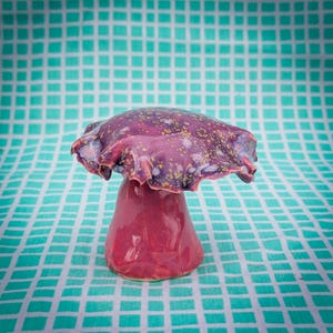 May include: A ceramic mushroom sculpture with a deep red stem and a ruffled, purple cap speckled with gold and white. The mushroom is set against a teal and white checkered background.