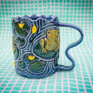 May include: A ceramic mug with a blue exterior, featuring a frog design. The mug has a wavy rim and a curved handle. The design includes lily pads and yellow flowers. The mug is approximately 4 inches tall.