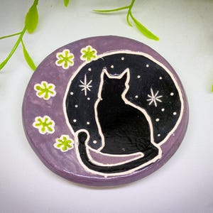 May include: A purple ceramic dish with a black silhouette of a cat sitting on a crescent moon. The moon is surrounded by white stars and green four-leaf clover designs.