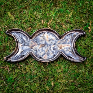 May include: A ceramic triple moon altar tray with a glossy, iridescent finish. The tray is shaped like three crescent moons connected together. The colors include shades of silver, blue, and brown. The tray is set against a background of green moss.