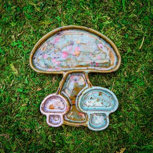 May include: A ceramic mushroom-shaped dish with two smaller mushroom-shaped bowls attached. The large mushroom is green, brown, and white with a speckled glaze. The smaller mushrooms are blue and white.