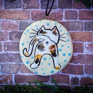 May include: A ceramic ornament featuring a hand-painted calico cat with a white, brown, and orange pattern. The cat is depicted with its back to the viewer, its tail curled around its body. The ornament is decorated with blue polka dots on a cream background.