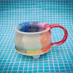 May include: A handmade ceramic mug with a colorful, abstract design. The mug features a white base with vertical stripes of blue, yellow, orange, and red. The handle is red. The mug sits on a small, white foot.