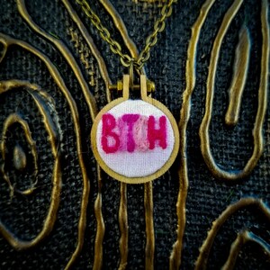May include: A pink and white embroidered pendant necklace with the word "BTCH" on it. The necklace is hanging from a gold chain on a black background.