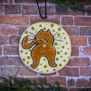 May include: A ceramic ornament shaped like a cat's rear end with a tail curled around its body. The ornament is painted in shades of brown and orange with green polka dots on a white background.