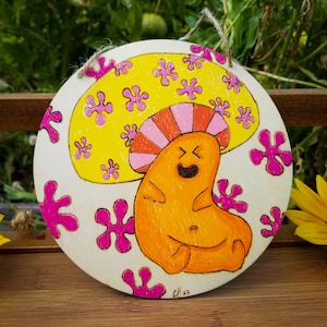 May include: A round wooden sign with a painted image of a smiling orange creature with a pink and orange mushroom hat. The background is white with pink and yellow flower-like shapes.