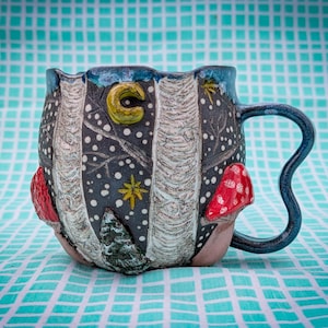 May include: A ceramic mug with a whimsical design. The mug features a dark blue background with white dots, a yellow crescent moon, and a red mushroom. The handle is a dark blue, curved shape.