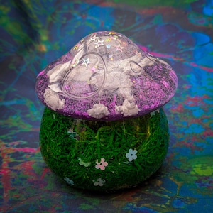 May include: A decorative mushroom-shaped container. The top is a translucent purple with embedded stars and white cloud-like shapes. The base is a green, moss-like texture with small white and pink flower accents. The background is a colorful, abstract pattern.