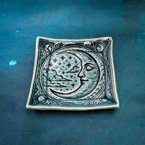 May include: A square ceramic trinket dish with a celestial design. The teal dish features a crescent moon with a serene face, surrounded by stars and clouds. Intricate patterns frame the moon, adding a touch of elegance to this decorative piece.