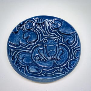 May include: A round, cobalt blue ceramic piece featuring a frog in a pond setting. The frog is centered, surrounded by lily pads and water lines. The design is carved into the ceramic, creating a textured, dimensional effect. The piece has a glossy finish.